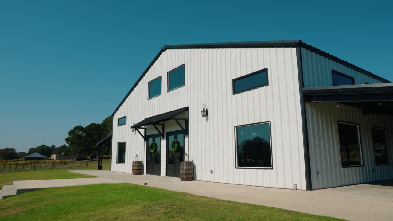 A side-angle shot of a charming wedding venue at a South Georgia vineyard. The modern farmhouse-style building features white siding, large windows, and rustic barrel decor by the entrance