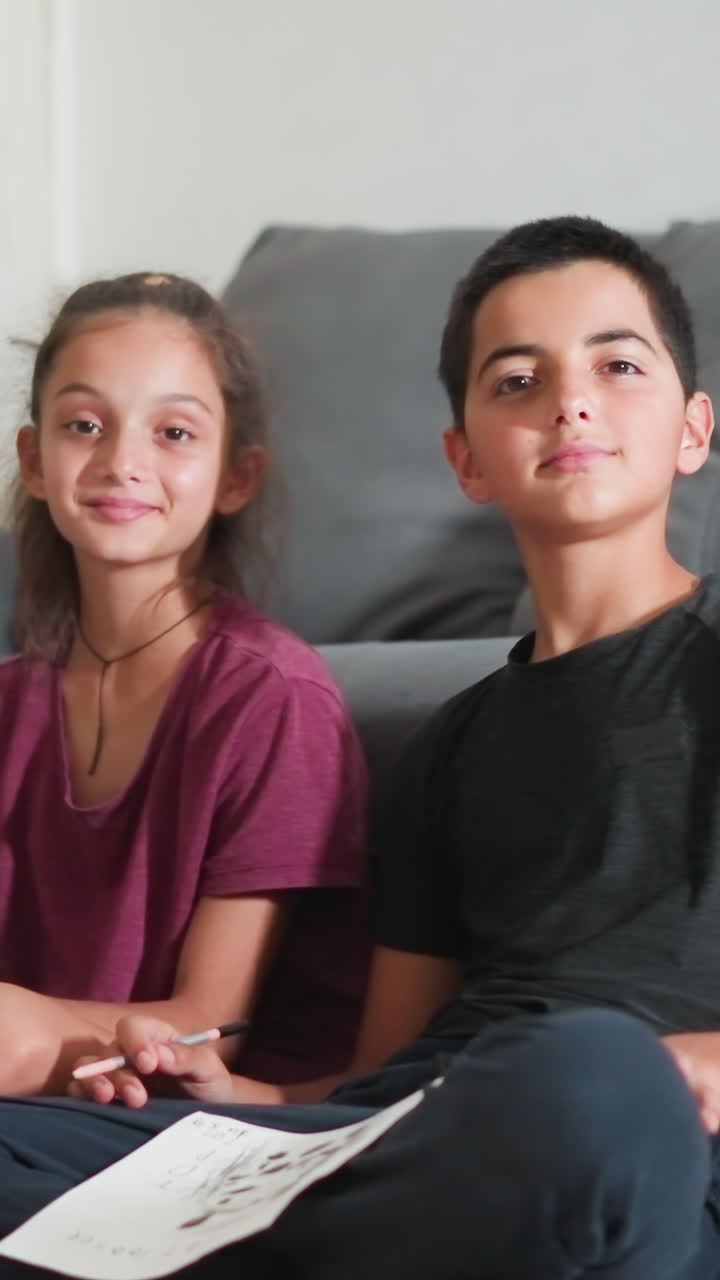 Hermanos adorables sentados cerca del sofá, sosteniendo trozos de papel y sonriendo a la cámara en una acogedora sala de estar bien iluminada, mostrando la alegría de un tiempo creativo juntos con blocs de dibujo y creando lazos a través del dibujo compartido.