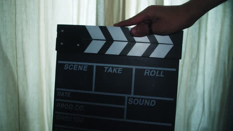 amazing slow motion shot of a young man using a clapperboard to start shooting a scene inside a room with curtains behind.