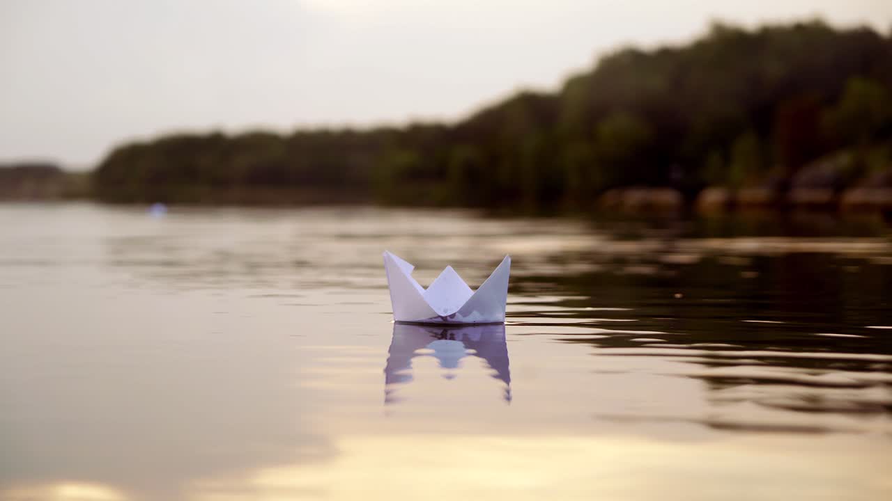 White paper boat is floating on the water near the forest background in the evening. Origami boat floats away into the distance along the river