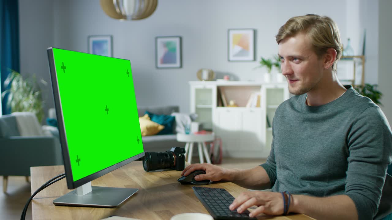 un bel giovane fotografo lavora su un computer personale con uno schermo verde mentre è seduto alla sua scrivania nell'accogliente appartamento.