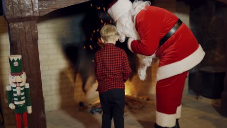 santa in red costume with children near the fireplace. santa claus shows little kids how to put wood into the fireplace inside the house.