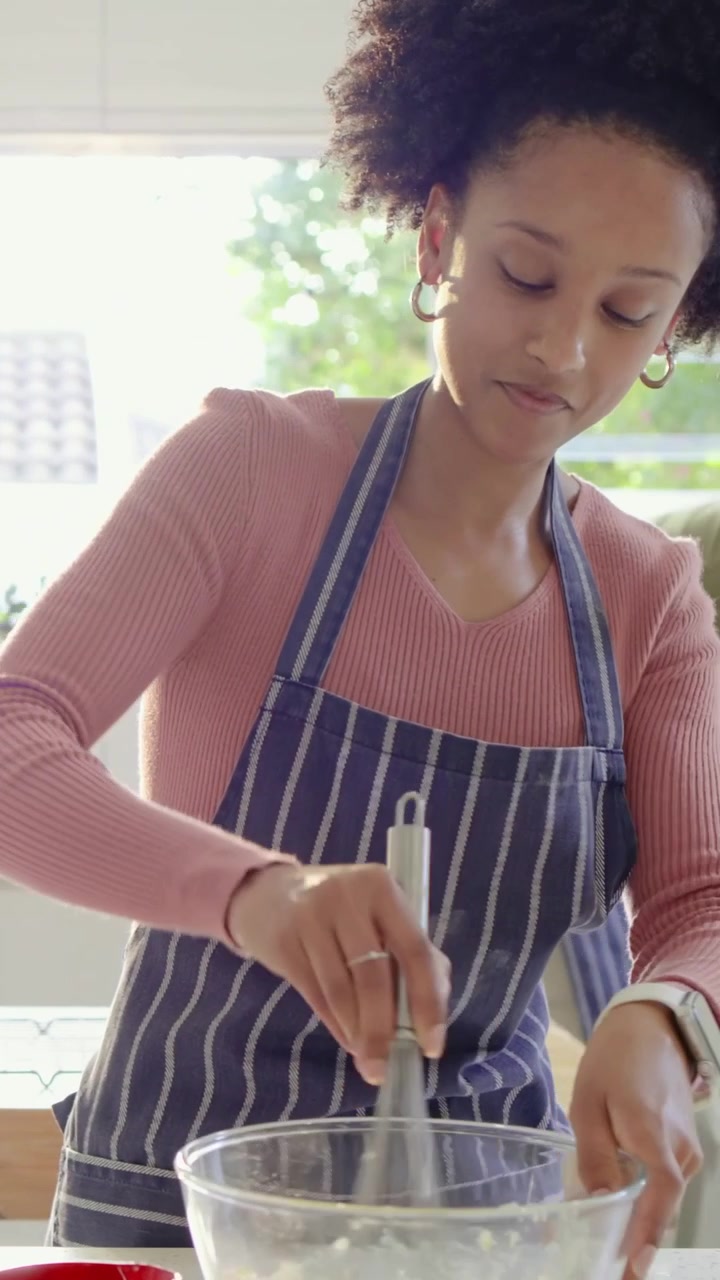 Video verticale: Donna che sbatte l'impasto in una ciotola in cucina, indossando un grembiule, mentre un uomo apre il frigorifero.