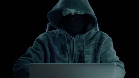 a man in hood sitting at table and coding on laptop. dark night. front view close up. hacker in balaclava mask working at computer. alpha channel chroma key transparent background.