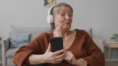 happy senior woman listening to music with headphones using smartphone while sitting in living room at home