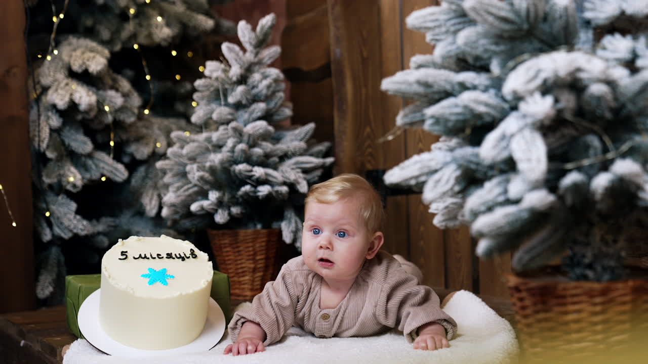Adorable blue-eyed infant with cute blond hair lies near the white cake. Lovely fir-trees around the kid.