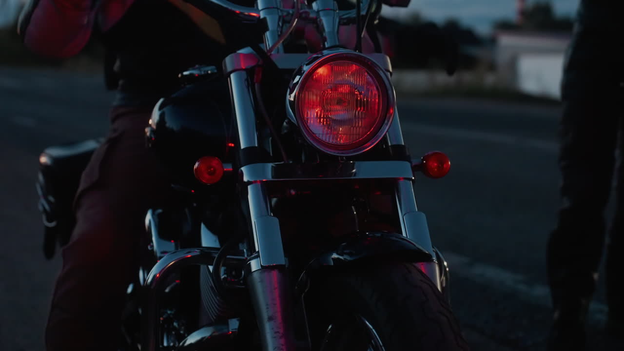 Motorcycle parked at roadside during dusk as passenger drops from seat, headlight casting strong beam across dim road surface, evening sky painted with fading light