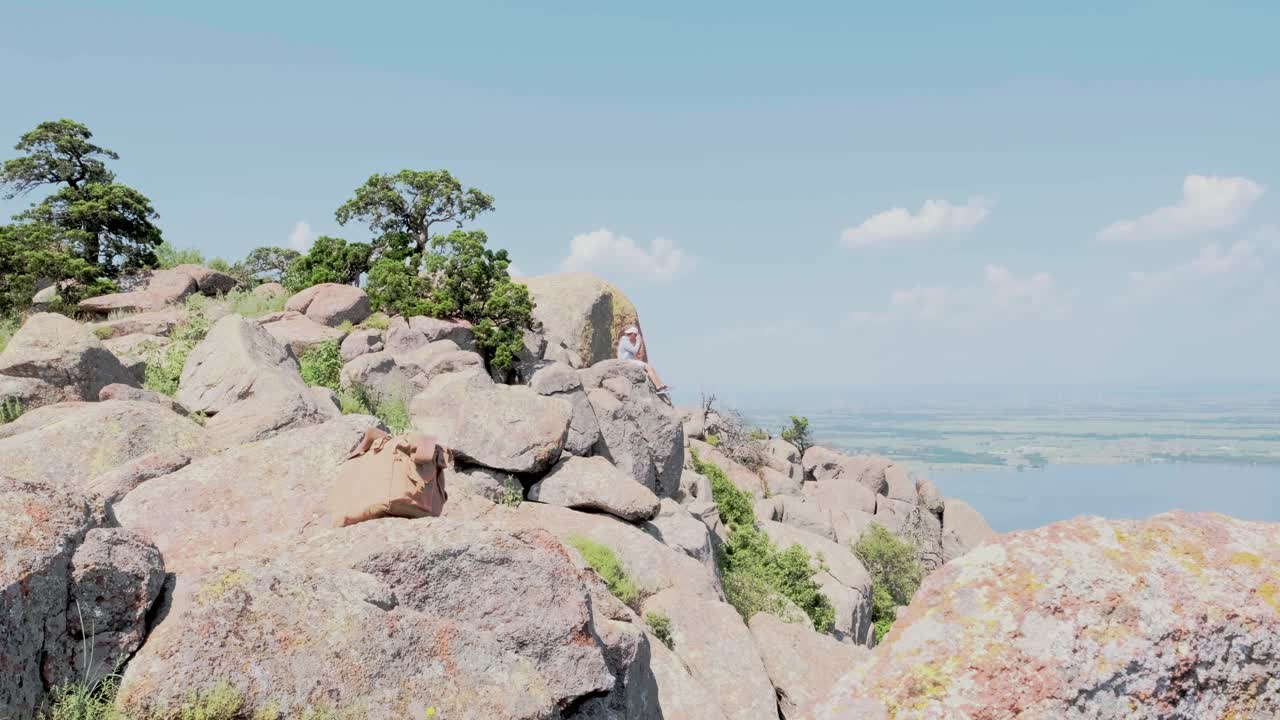 A person sitting on a rocky mountain overlooking a lake and wind turbines