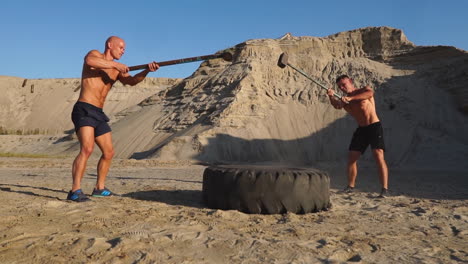 dos atletas masculinos entrenando juntos golpearon la rueda con un martillo al atardecer en las montañas en la arena. entrenamiento de resistencia