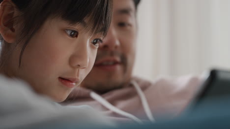 little-asian-girl-using-tablet-computer-watching-entertainment-with-parents-playing-game-on-touchscreen-device-browsing-online-relaxing-on-sofa-together-at-home-4k