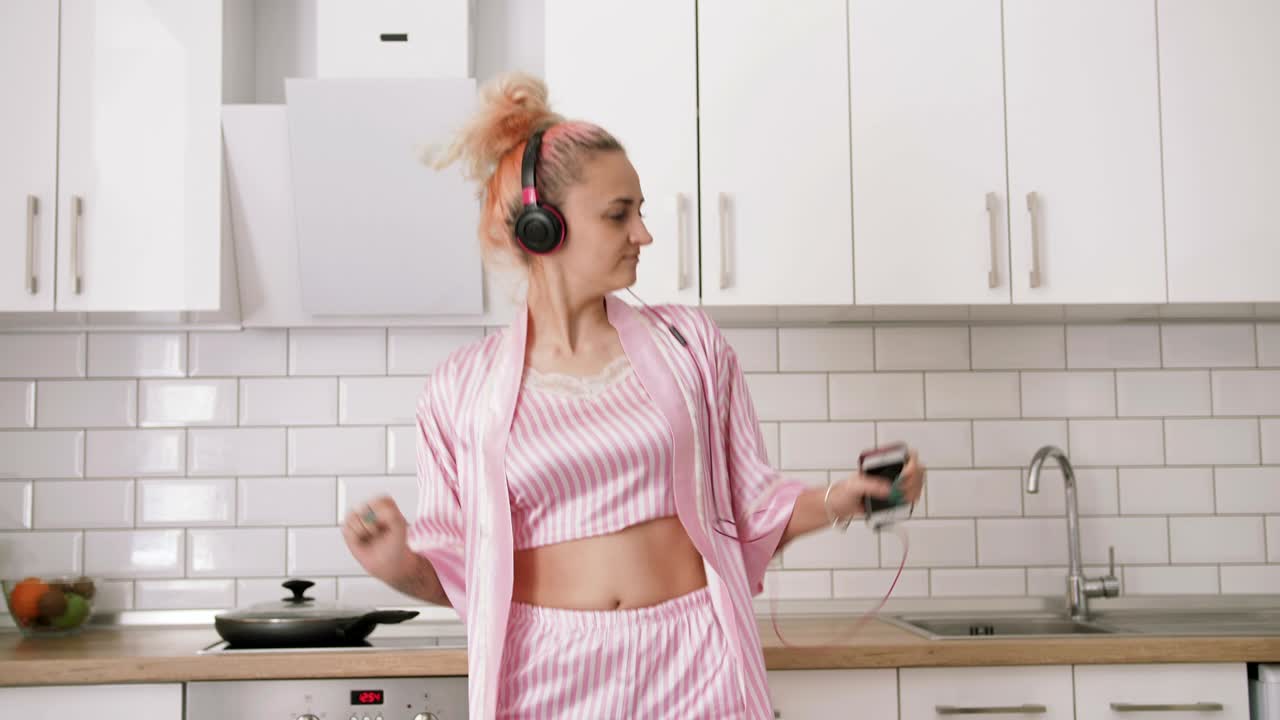 Giovane donna felice con i capelli rosa che balla in cucina, indossando vestiti rosa e ascoltando musica con le cuffie