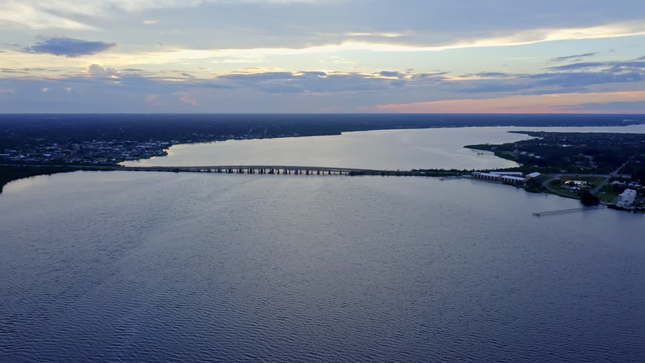 A sweeping aerial view shows a bridge cutting across a vast river as twilight colors paint the horizon, blending calm water, distant land, and sky in quiet evening balance