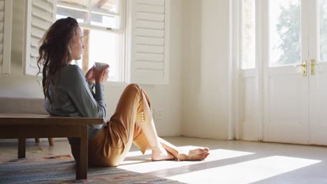 Thoughtful-caucasian-woman-sitting-on-floor-in-sunny-cottage-drinking-coffee,-looking-out-of-window