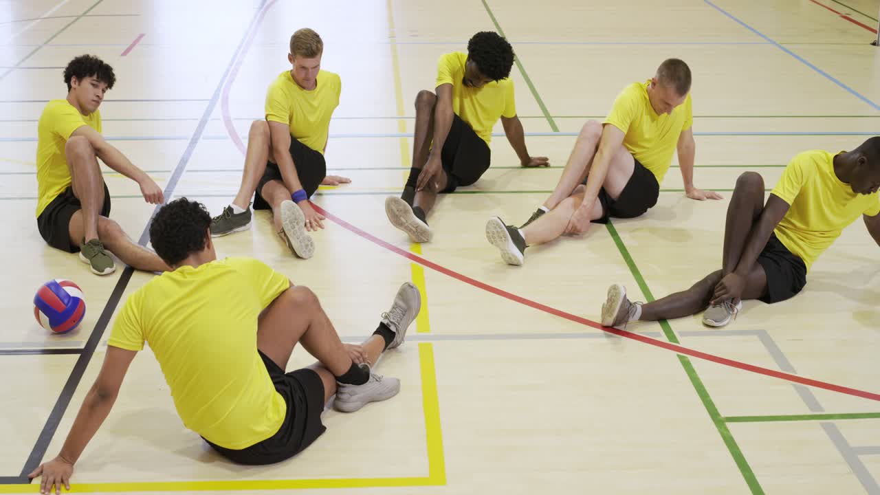 Sechs erwachsene Teamkollegen in gelben Trikots führen in einer Sporthalle eine Schmetterlingsdehnung durch, ein Volleyball liegt daneben.