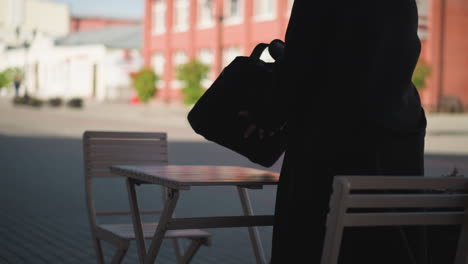 back view of woman placing her bag on wooden table and sitting down at outdoor cafe with urban building and someone walking in blurred background