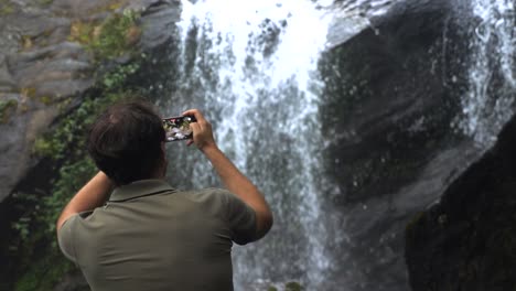 travel man with mobile phone shooting video of waterfall in dark rainforest