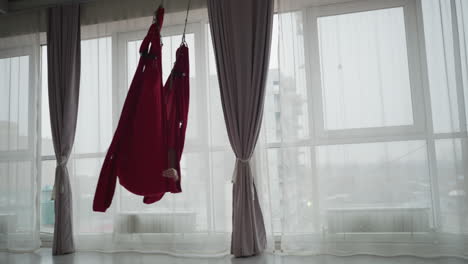 bright yoga studio with floor to ceiling windows and sheer curtains, flexible woman lying comfortably on red silk hammock swinging gently, conveying calm focus balance and wellness practice