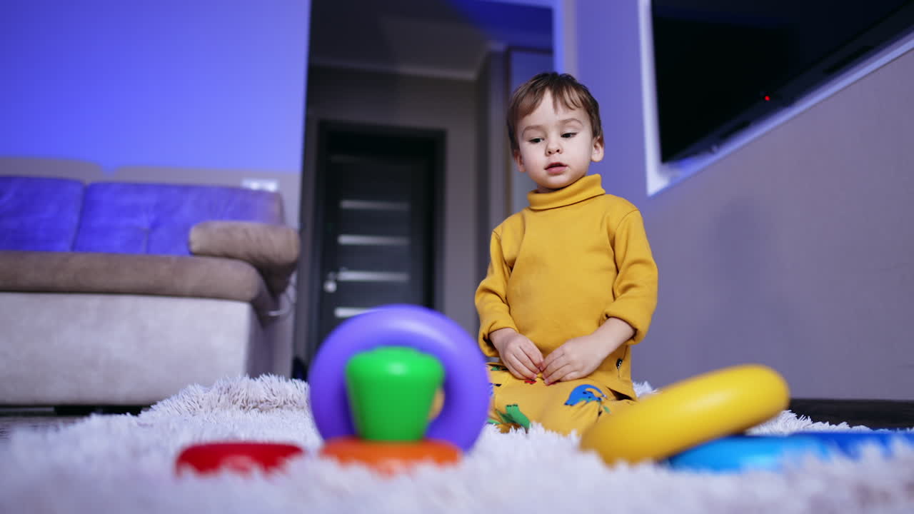 Funny boy in yellow clothes sits on the carpet. Kid plays with rings from toy pyramid. Low angle view.