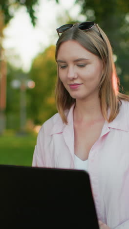 mujer con camiseta rosa con gafas apoyadas en la cabeza sonríe cálidamente mientras trabaja en su computadora portátil al aire libre, la luz del sol crea un brillo suave en ella, reflejando un momento de paz