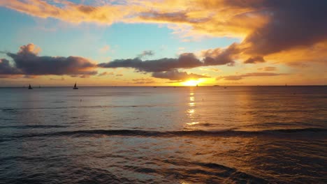 aerial drone flying over beautiful ocean sunset with sailboats on waikiki beach in honolulu, hawaii at golden hour with colorful orange sky as waves crash into breakwall with