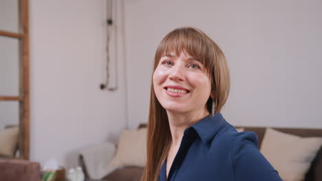 young girl seated on sofa smiling gently with hands lifted mid-gesture, wearing navy outfit in cozy modern living room with decorative pillows, wooden mirror, table clock, and soft ambient lighting