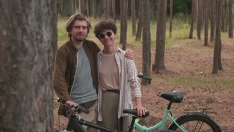 joven afectuoso abrazando a su feliz esposa con gafas de sol mientras ambos están de pie con sus bicicletas en un entorno rural