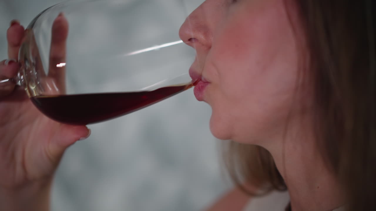 Close up side view of elegant young woman sipping red wine from large glass, soft lighting highlighting facial features, indoor setting with blurred geometric background