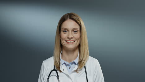 portrait of the young beautiful cheerful woman doctor smiling and looking at the camera while standing on the gray background