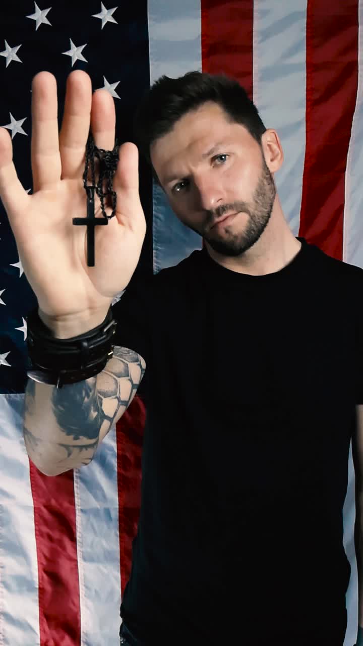 Patriotic man lifts up a holy black colored cross on a chain in his hands and shows it to the camera in front of a vertically hanging US American United States flag that is slowly moving in the wind