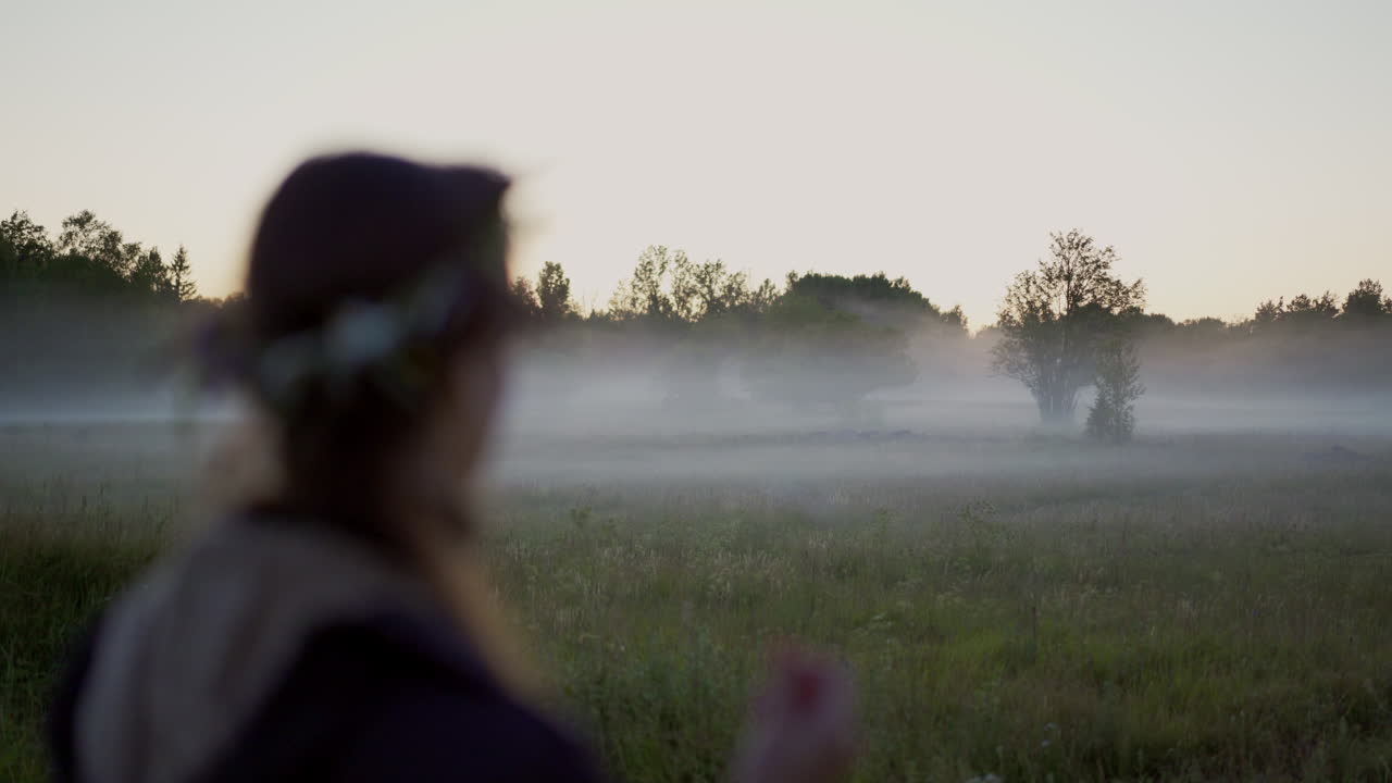 A woman wearing a flower wreath stands at a fog-covered grass field, gazing off into the distance on a serene midsummer evening. Tranquility of the Nordic nature surroundings and peaceful atmosphere.