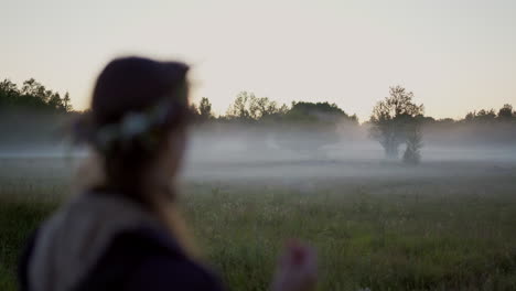 a woman wearing a flower wreath stands at a fog-covered grass field, gazing off into the distance on a serene midsummer evening. tranquility of the nordic nature surroundings and peaceful atmosphere.