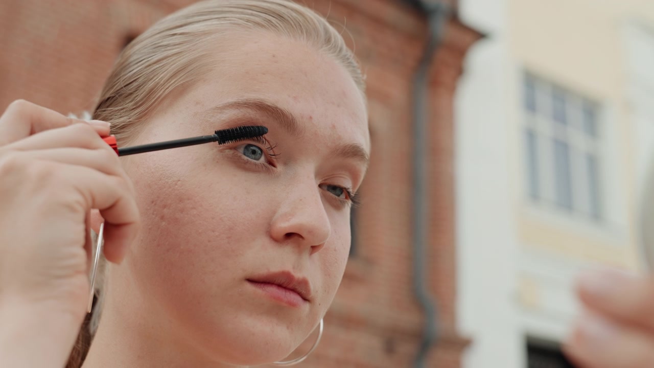 Young woman receives makeup application outdoors focusing on eye and face details. hand applies mascara near her ear while another holds compact mirror with red accents. background features