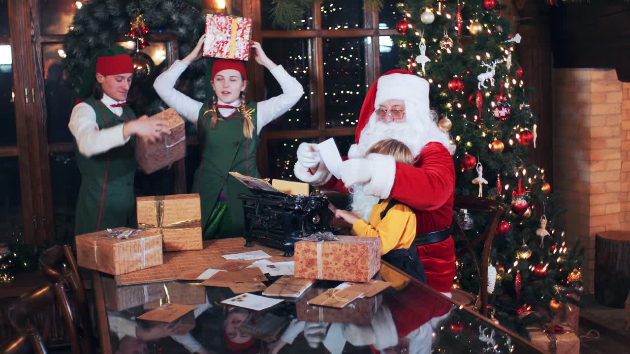 Elves collect gifts for holiday packages for children for the new year. Santa Claus with a boy sitting at a table typing letters on a typewriter