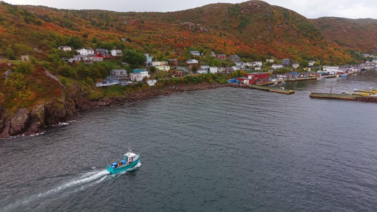 Cinematic aerial footage captures a boat gliding through the tranquil waters of Petty Harbour, Newfoundland, surrounded by autumn-colored hills and a quaint coastal village under a crisp fall sky