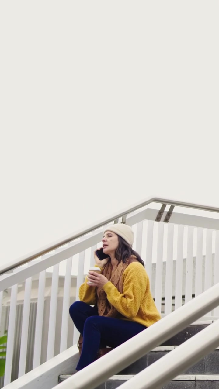 Video verticale: Donna in maglione giallo che risponde a una chiamata, tiene il telefono, tazza sulle scale, spazio per il testo.