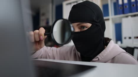 un ladrón con una máscara y buscando información usando una lupa en la computadora de la oficina