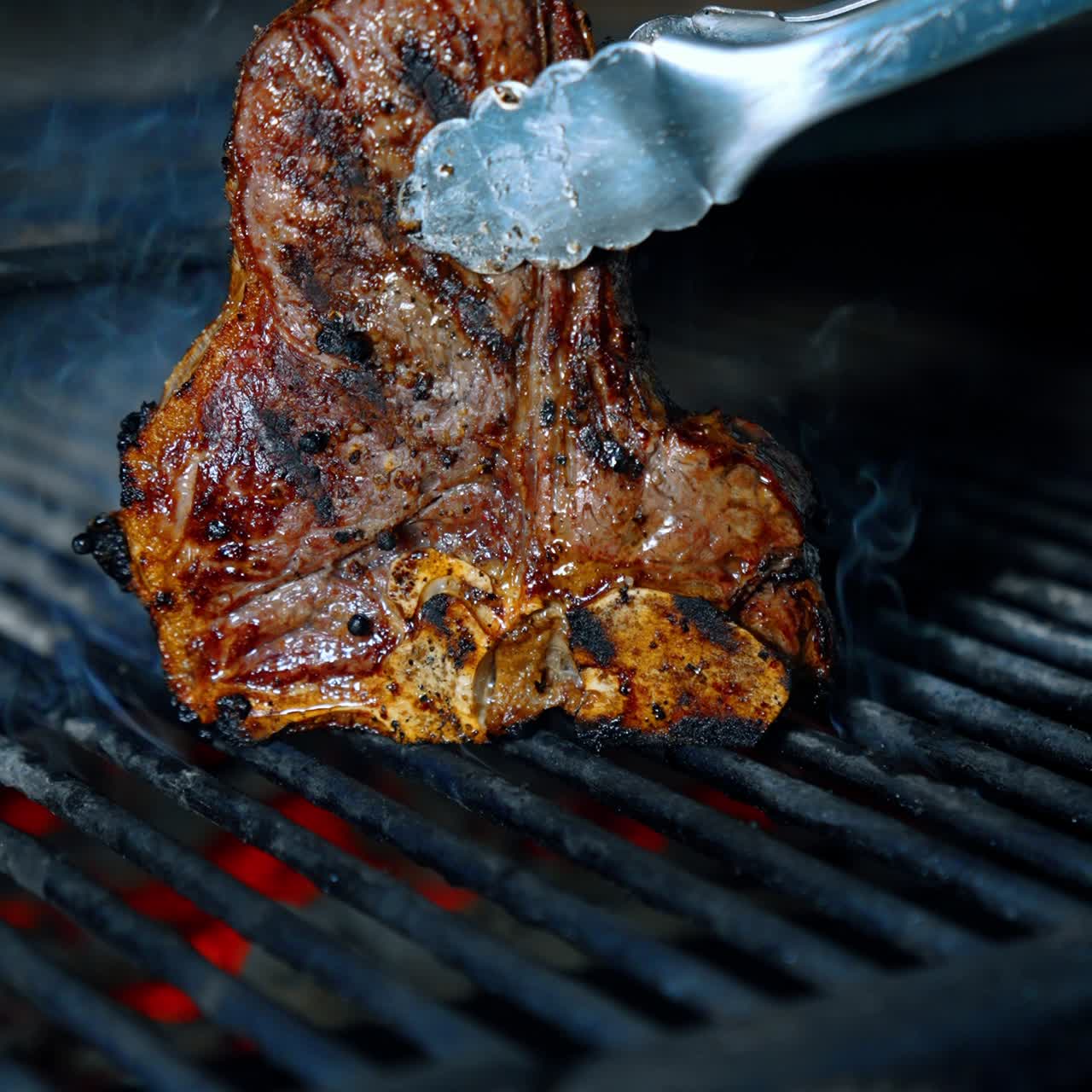 Piece of beef on the bone is cooked on the grill. Meat is turned with the tongs. Close up. Vertical view