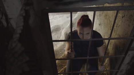 young woman on her knees raising her eyes and looking outside a cage's rusty bars