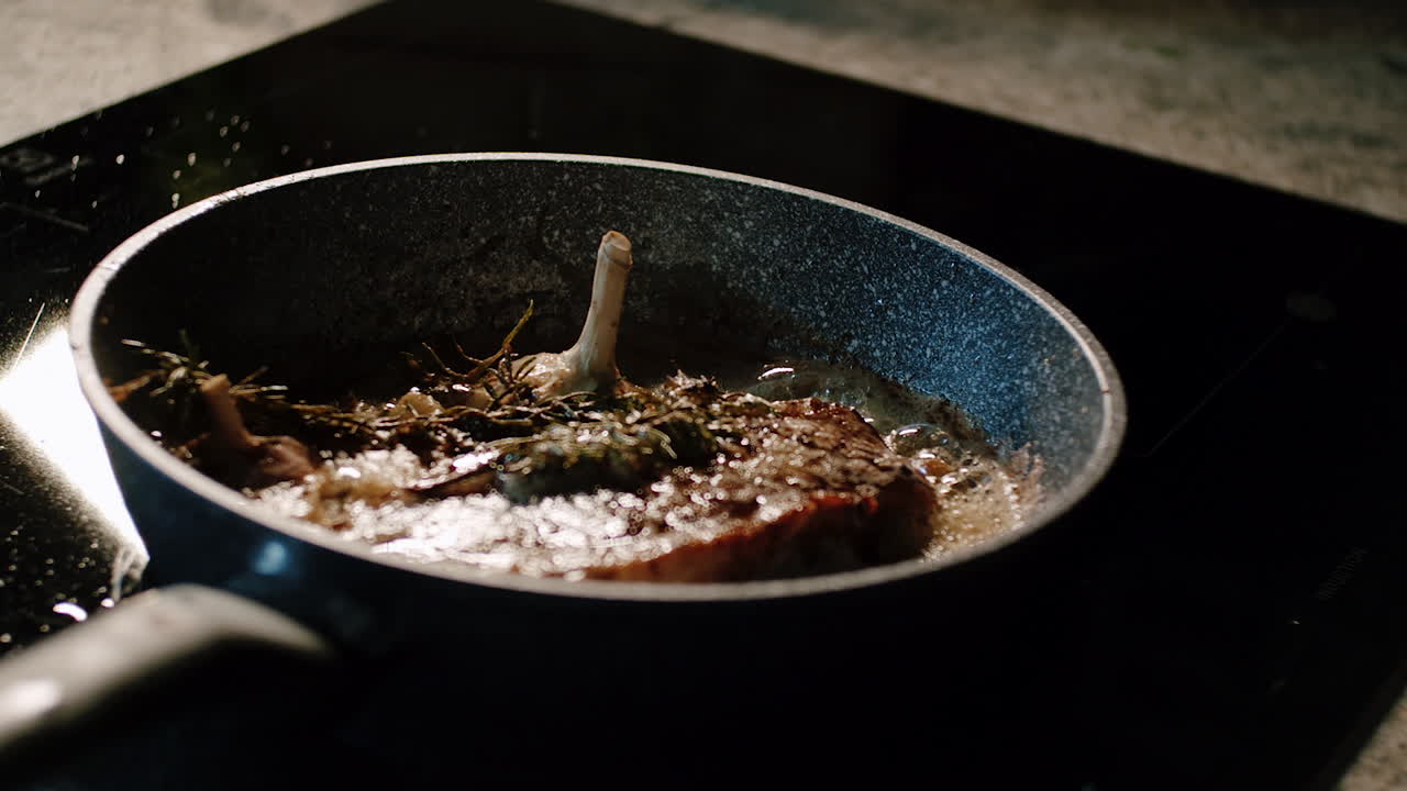 Cottura di bistecca con aglio e rosmarino in padella su piano cottura in ceramica.