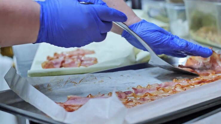 Blue gloves arrange bacon slices on lined trays inside commercial kitchen space