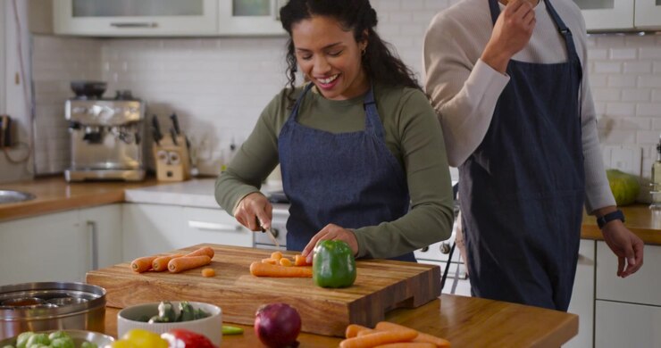 Coppia afroamericana che taglia in cucina, donna che affetta una carota su un tagliere, uomo che si avvicina e assaggia.