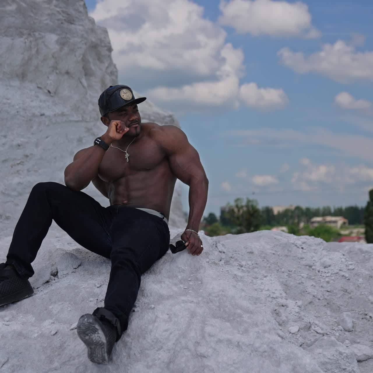 Full length view of the athletic fit model with naked torso sitting at the sand. Strong handsome man posing on blue sky background