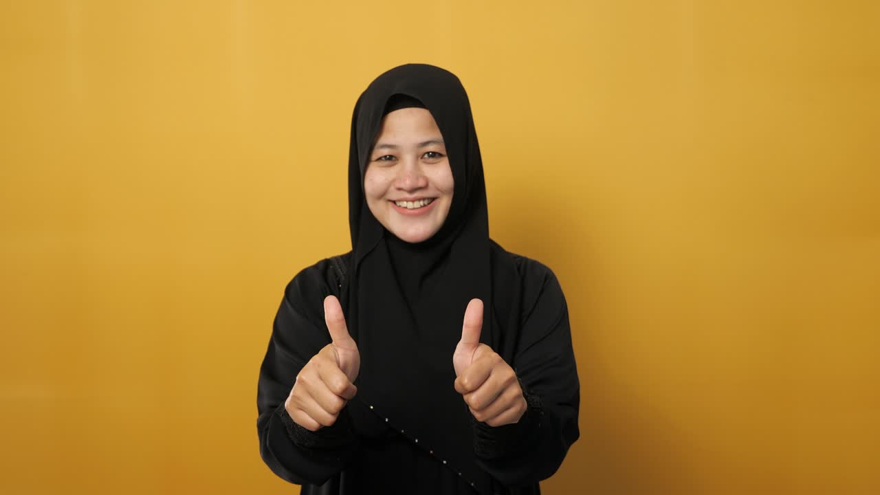히자브를 입은 젊은 아시아 무슬림 여성의 초상화는 엄지손가락을 올려 놓은 몸짓, 웃는 행복한 얼굴, 승인된 ok 표지, 노란색에 대항합니다.