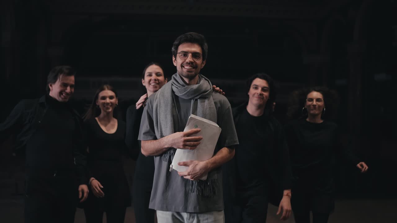 retrato de un hombre moreno confiado con gafas redondas director de escenario en una camiseta gris y bufanda a quien los actores en trajes negros corren y abrazan por detrás en el escenario en el teatro