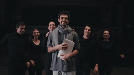 Retrato-De-Un-Hombre-Moreno-Seguro-De-Sí-Mismo-Con-Gafas-Redondas,-Director-De-Escena-Con-Una-Camiseta-Gris-Y-Una-Bufanda,-Al-Que-Los-Actores-Con-Trajes-Negros-Se-Acercan-Corriendo-Y-Abrazan-Por-Detrás-En-El-Escenario-Del-Teatro.