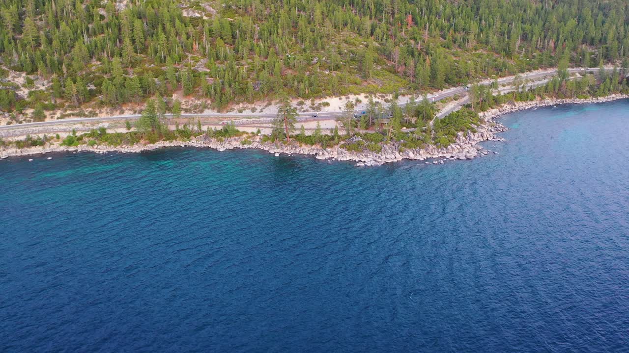 Lakeside Road with Cars Driving, Pine Tree Forest, Rocky Riprap Bank and Clear Blue Water Along Scenic Coast At Lake Tahoe California - aerial drone flyover