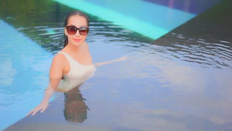 Hermosa-Mujer-Asiática-Dentro-De-Una-Piscina-De-Lujo-Futurista-Con-Gafas-De-Sol-Y-Monokini-Blanco-De-Un-Hombro-Mirando-La-Lente-De-La-Cámara-Y-Apoyándose-En-El-Borde-De-La-Piscina