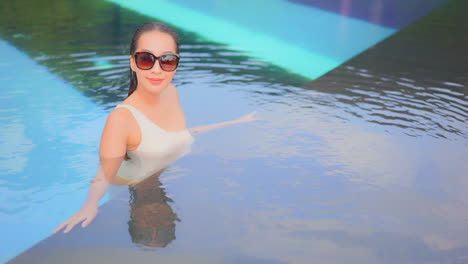 beautiful asian woman inside futuristic luxury swimming pool wearing sunglasses and white one-shoulder monokini looking at camera lense and lean on the edge of the pool