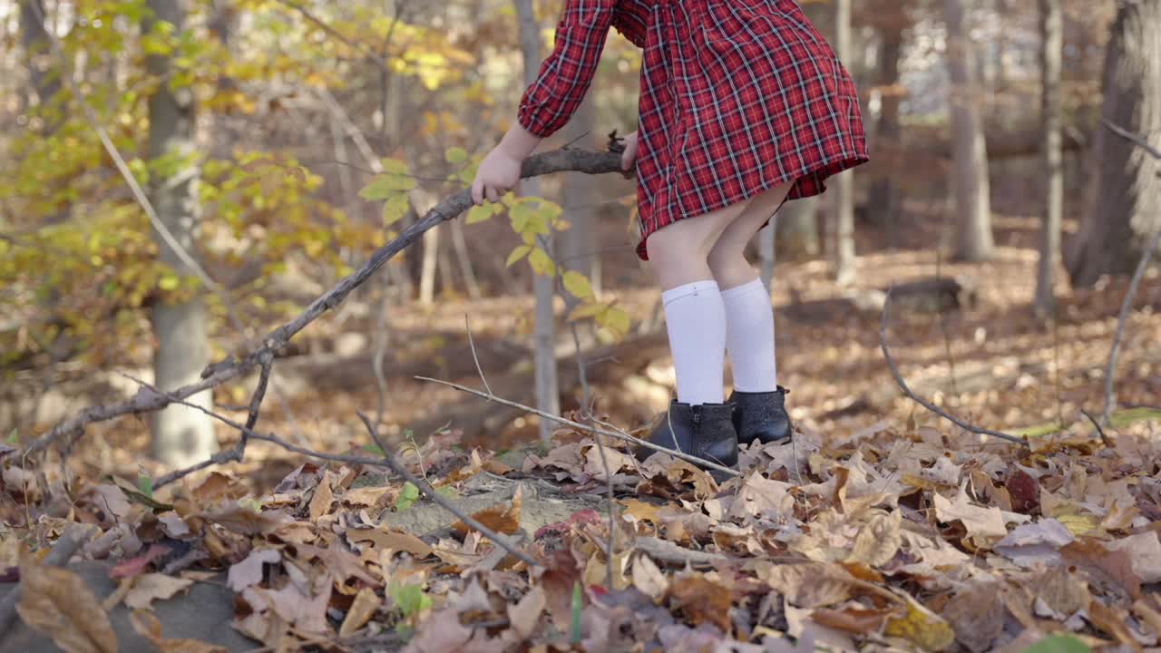 Mädchen im roten Kleid spielt im Wald, umgeben von Herbstblättern, in Zeitlupe bei Sonnenuntergang, während sie mit einem langen Ast gefallenes Laub zusammenharkt.