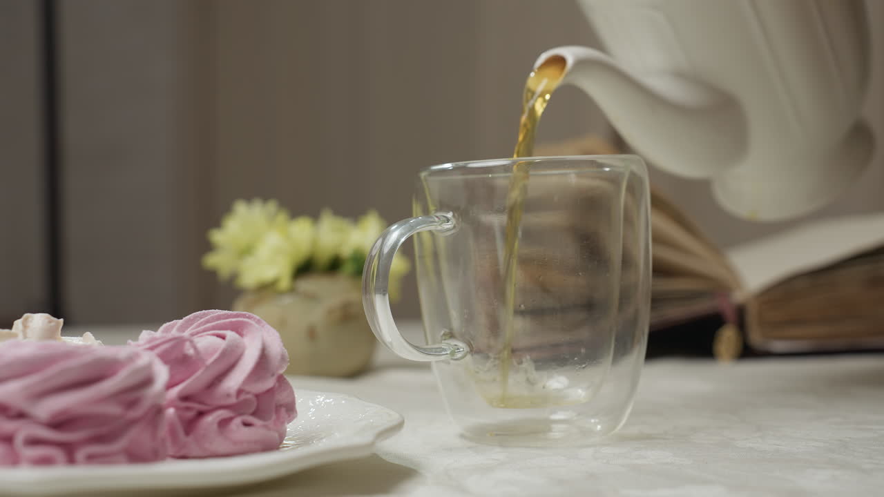 Close up hand view of person gently pouring herbal tea from white ceramic teapot into clear glass cup beside plate of pink meringue dessert and vase of yellow flowers on cozy kitchen table